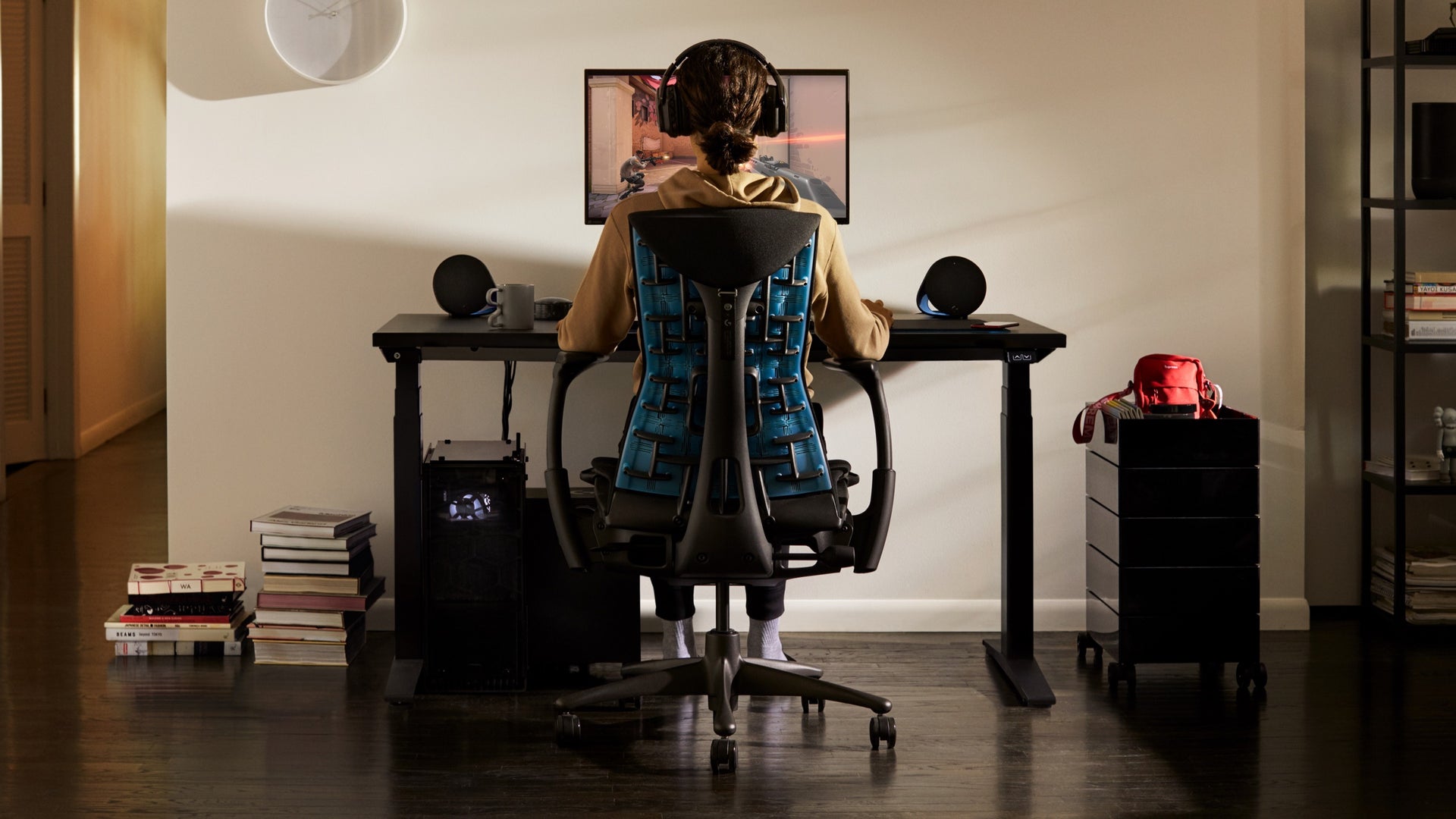家居环境里，一个人坐在Embody电竞椅上，看着电竞桌上的Ollin显示器挂臂连接的显示器。